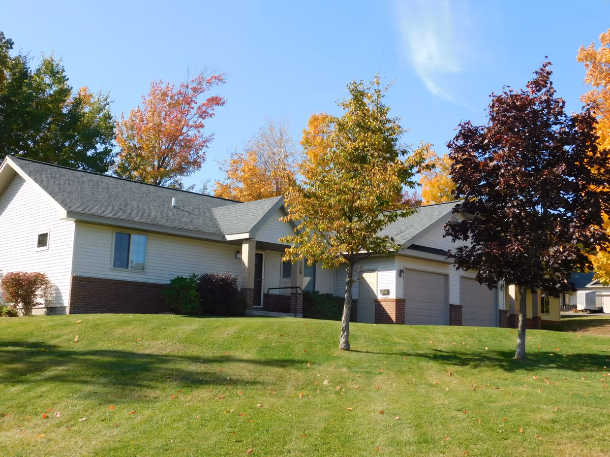 Single-story light-colored residential building with attached garages, a grassy front lawn and autumn trees under a clear blue sky.