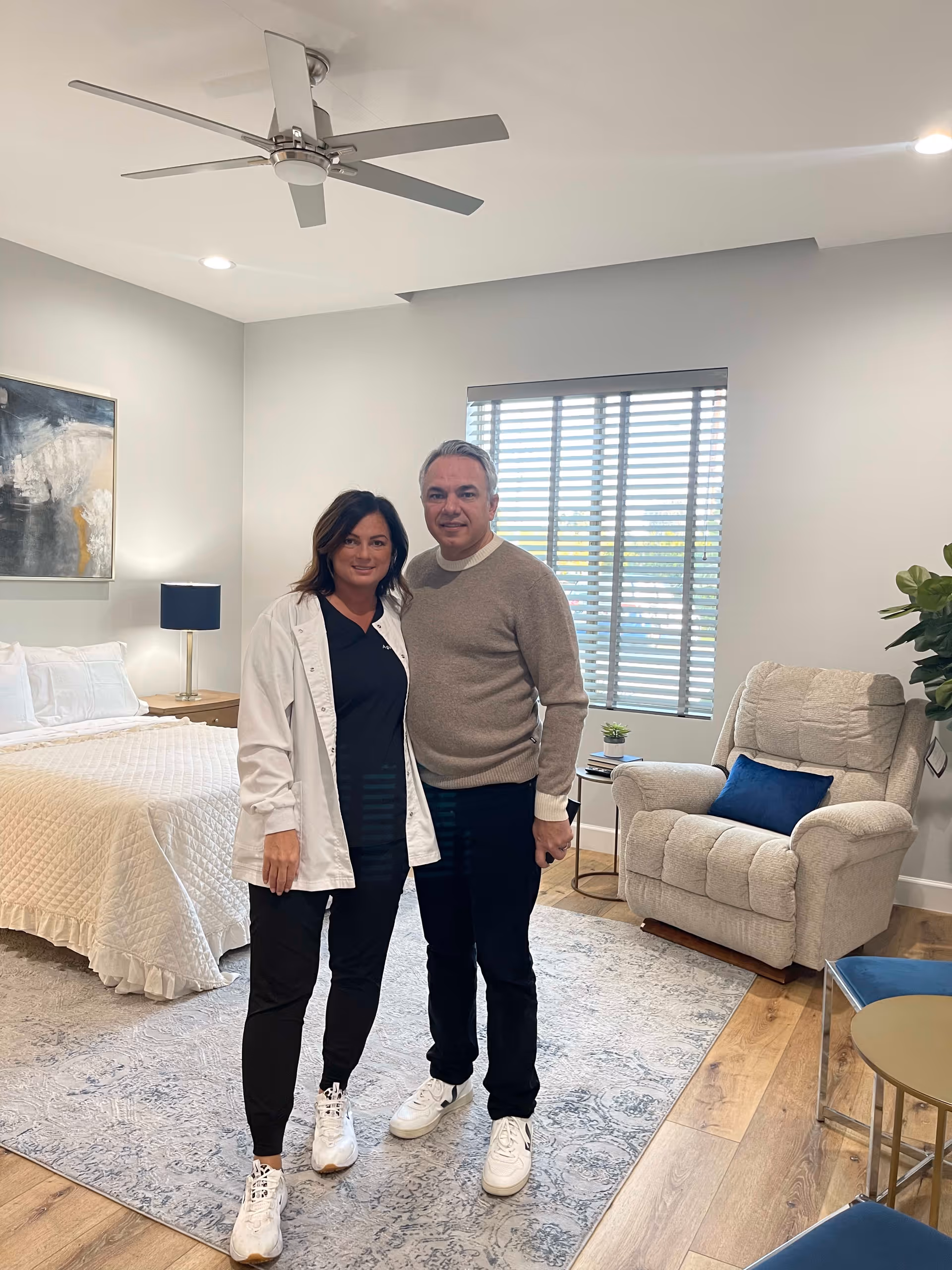 A man and a woman standing together in a well-lit bedroom. The room features a bed with a white quilted bedspread, a bedside table with a lamp, a large window with blinds, a beige recliner chair with a blue pillow, a small round side table with a plant and books, and a ceiling fan. The floor is wooden with a patterned area rug.