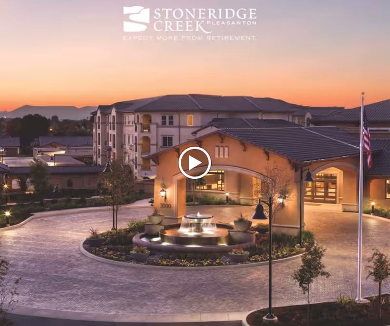 Exterior view of Stoneridge Creek Pleasanton senior living facility at sunset, featuring a circular driveway with a central fountain, landscaped plants, and an American flag on a flagpole.