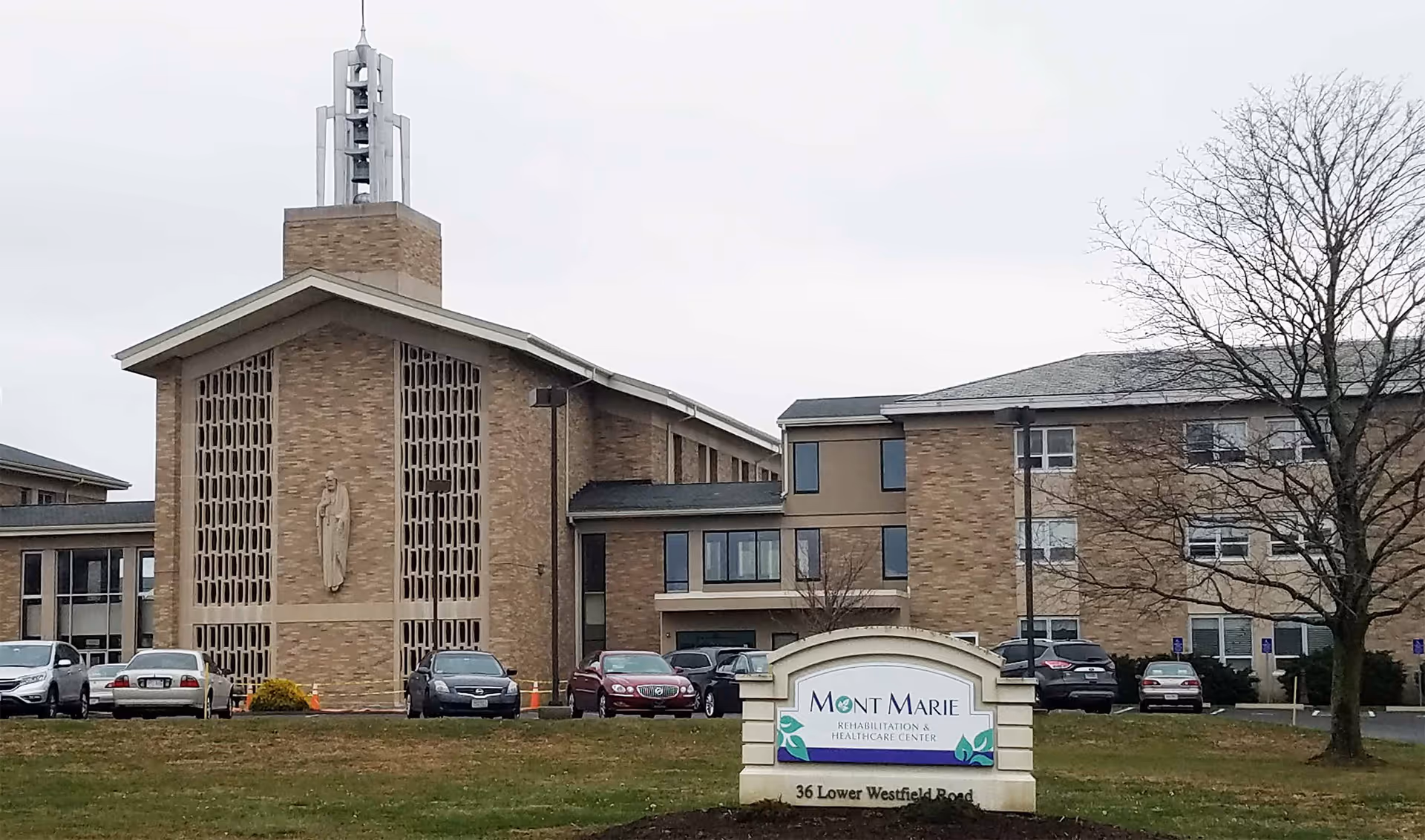 Front exterior of Mont Marie Rehabilitation & Healthcare Center with its entrance sign, parked cars, and a leafless tree on a cloudy day.