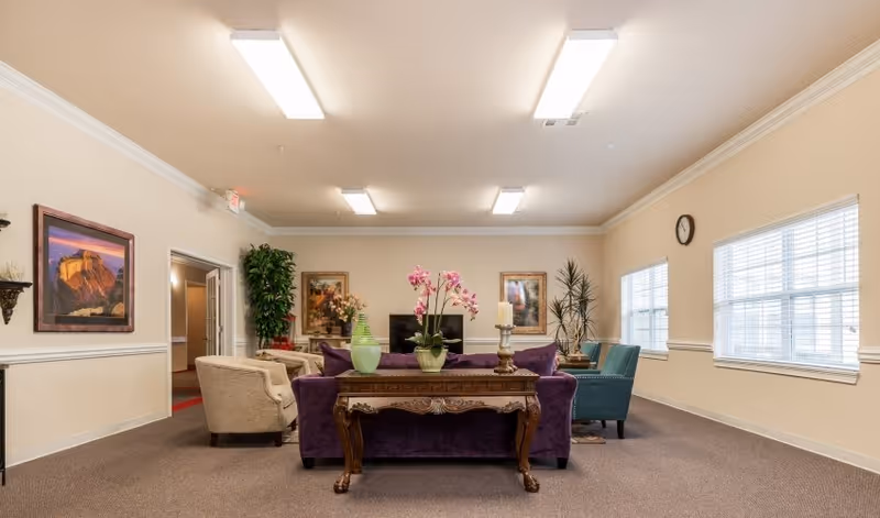 Communal lounge in a senior living facility featuring a purple sofa, armchairs, a decorative table with flowers and vases, framed artwork, and large windows.