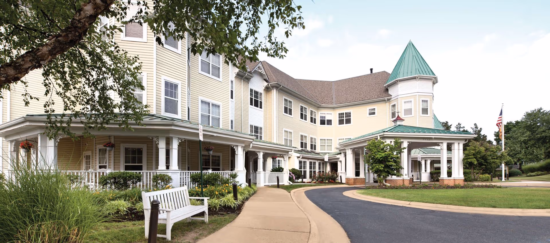 Exterior view of a senior living facility building with beige siding, white trim, and green metal roofs. The building features a covered entrance with white columns, a curved driveway, landscaped greenery, and a white bench near the sidewalk. An American flag is visible in the background.