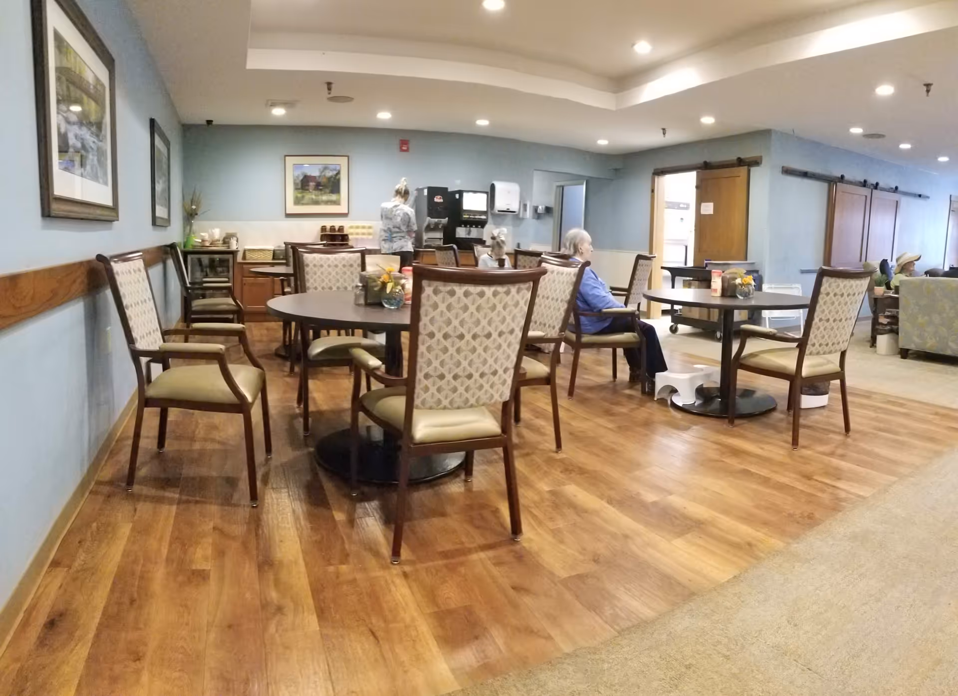 A dining area in a senior living facility with round tables and cushioned chairs. Several elderly residents are seated at the tables, and a staff member is standing near a beverage dispenser against the wall. The room has wooden flooring, light blue walls, framed artwork, and recessed ceiling lights.