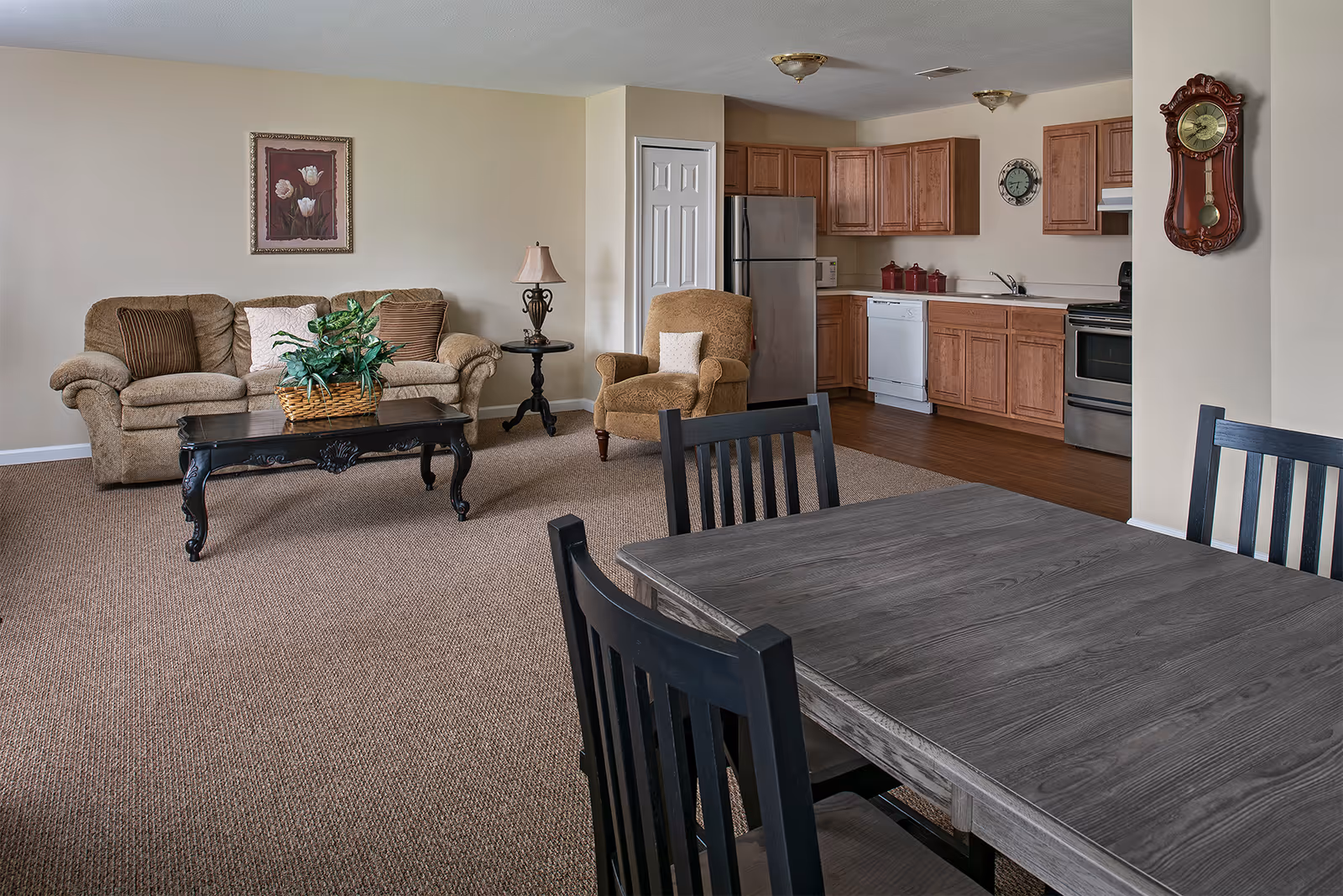 A cozy living area with a beige sofa, an armchair, a black coffee table with a plant, and a side table with a lamp. The room opens into a kitchen with wooden cabinets, stainless steel refrigerator, dishwasher, and stove. A dining table with black chairs is in the foreground. The walls are decorated with a framed floral picture and a wall clock.