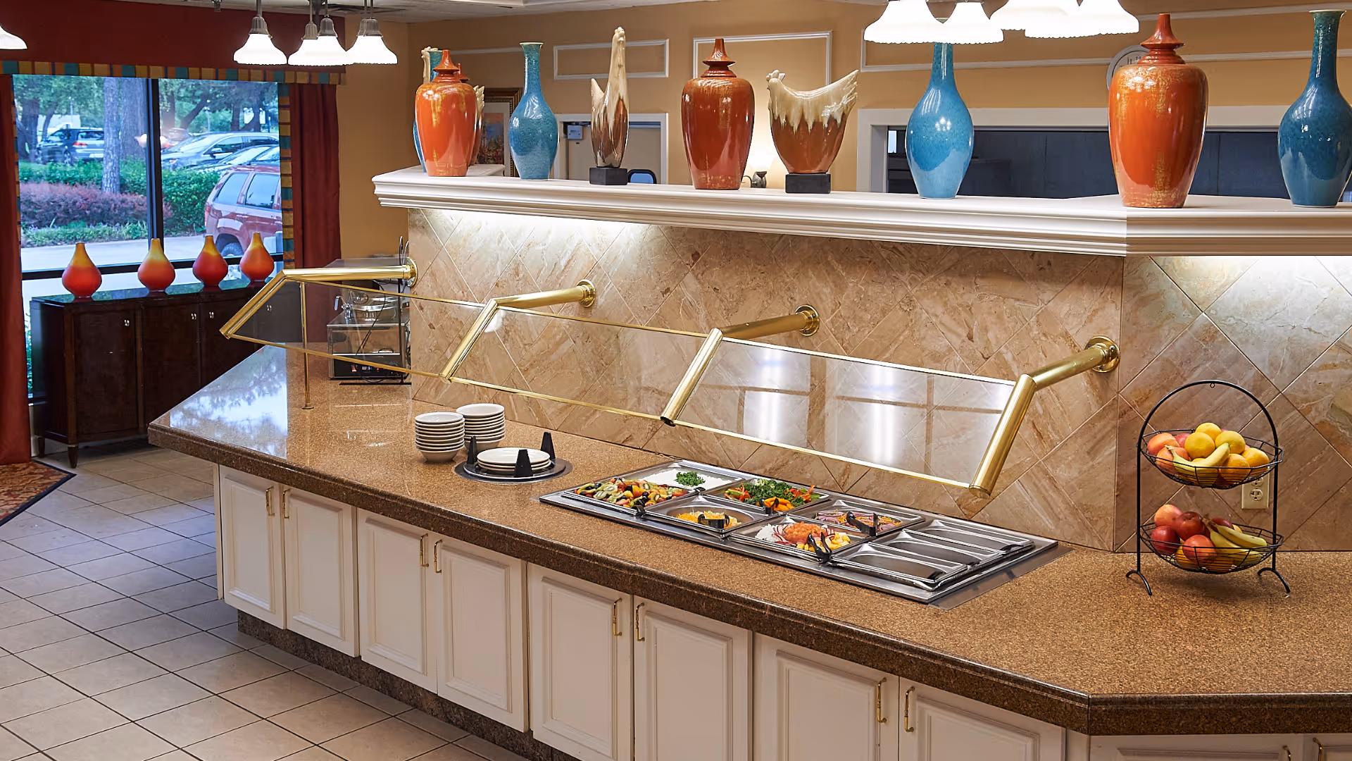 Interior view of a buffet counter with a glass sneeze guard, featuring trays of food, stacks of plates, and a two-tiered fruit basket with apples and bananas. Decorative vases are placed on the ledge above the counter, and large windows show a parking lot and greenery outside.