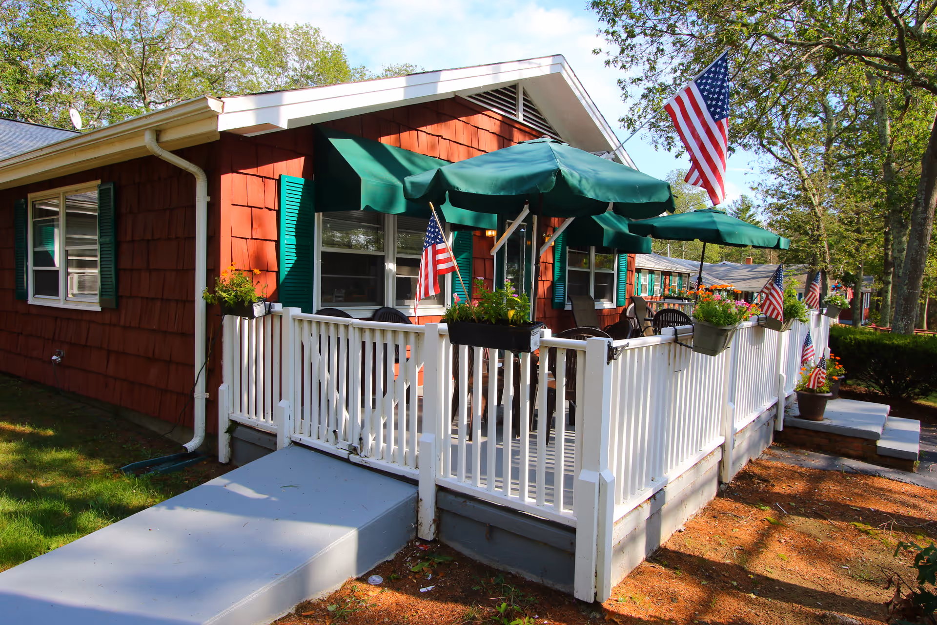 Exterior view of a red building with green shutters and a white fenced porch. The porch has green umbrellas, chairs, flower pots, and several American flags. Trees and greenery surround the area under a partly cloudy sky.