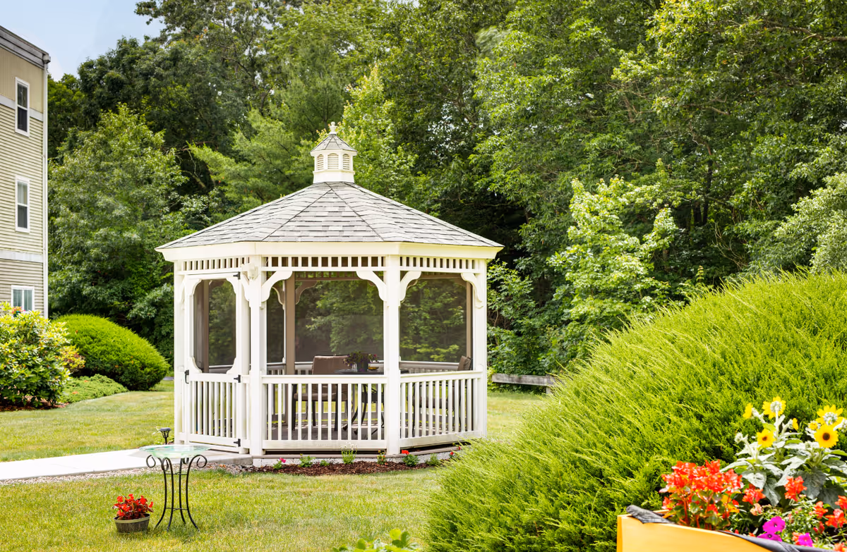 A white gazebo with a shingled roof sits in a green garden area surrounded by lush trees and bushes. There is a small table with chairs inside the gazebo, and colorful flowers and greenery are visible in the foreground. Part of a building is visible on the left side of the image.