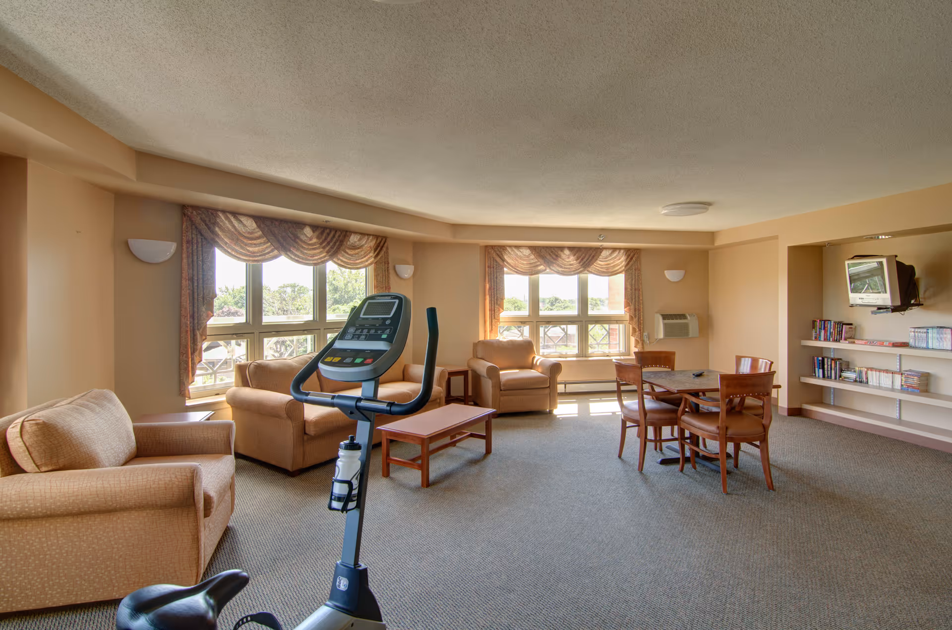 A spacious common area in a senior living facility featuring beige armchairs, a wooden coffee table, a round wooden table with four chairs, an exercise bike, large windows with patterned curtains letting in natural light, and a wall-mounted TV with shelves holding books and DVDs.