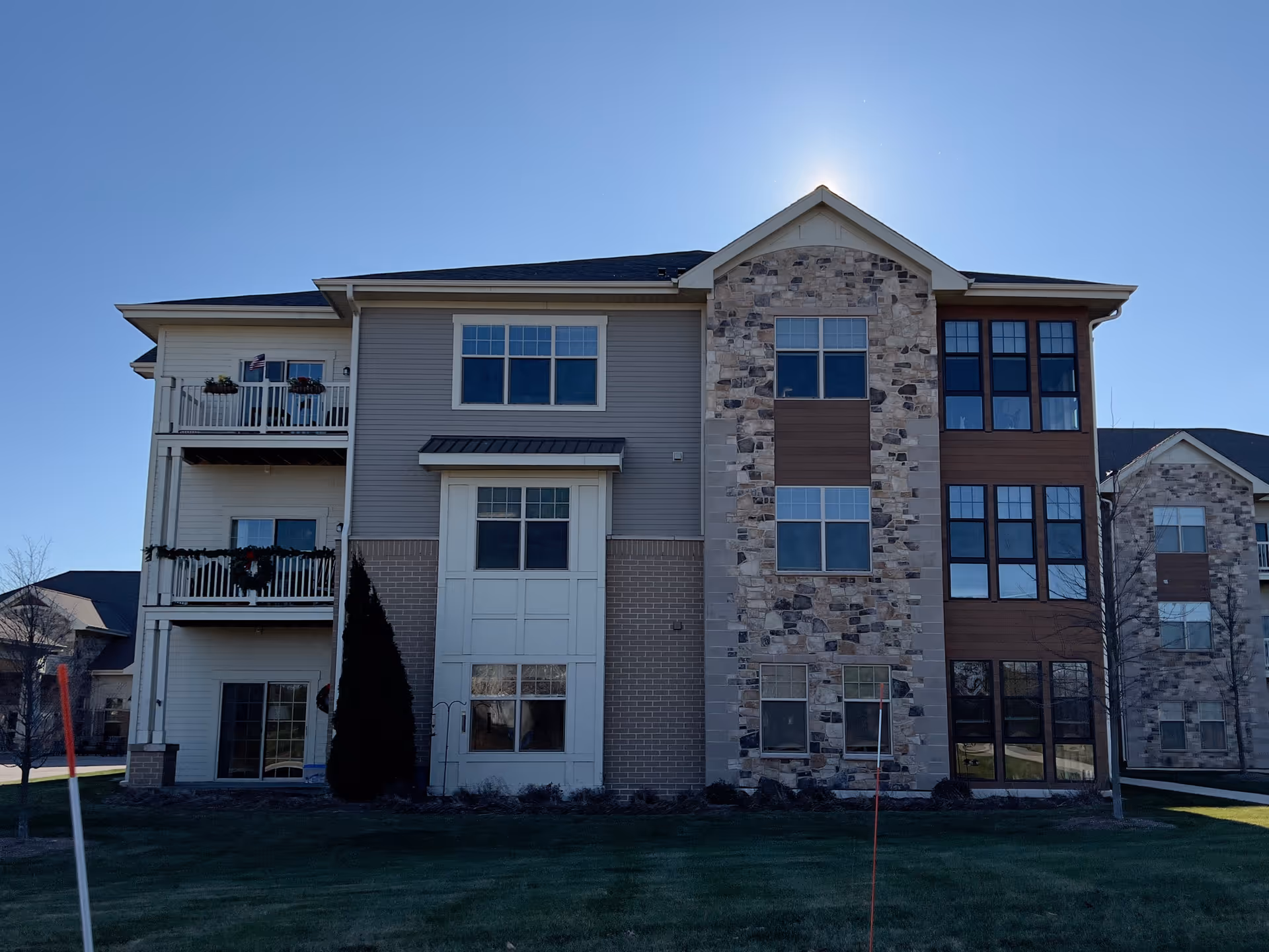 Exterior view of a multi-story residential building with a mix of stone, brick, and siding on the facade. The building has multiple windows and balconies, with some holiday decorations visible on the railings. The sun is shining behind the roof peak, and there is a well-maintained lawn in front.