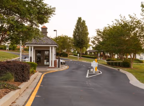 Entrance road to a senior living facility with a small guardhouse or gatehouse on the left side, surrounded by trees and landscaped greenery under an overcast sky.