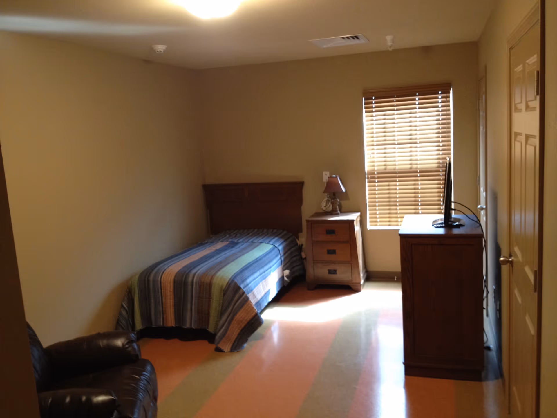 A small bedroom with a single bed covered with a striped blanket, a wooden nightstand with a lamp, a window with closed blinds, a wooden dresser with a TV on top, and a leather armchair in the corner.