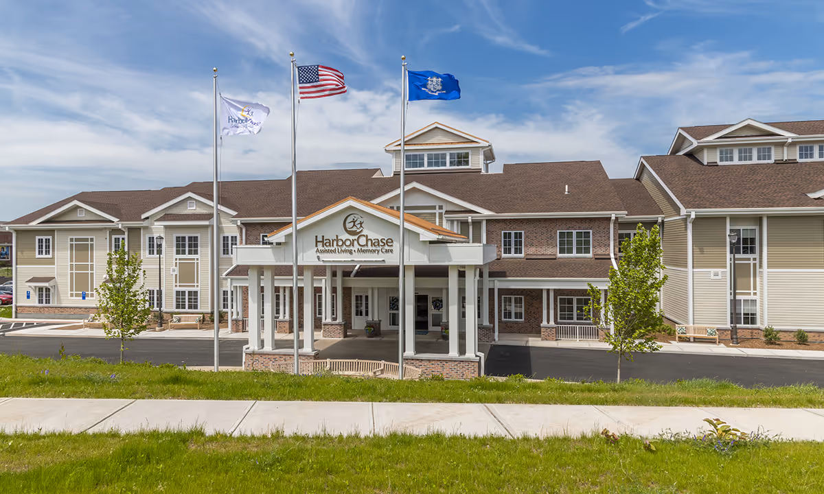Front exterior view of a large assisted living and memory care facility named HarborChase, featuring a covered entrance with three flagpoles flying the American flag, a state flag, and a facility flag. The building has a combination of beige siding and brick with multiple windows and a well-maintained lawn in front.