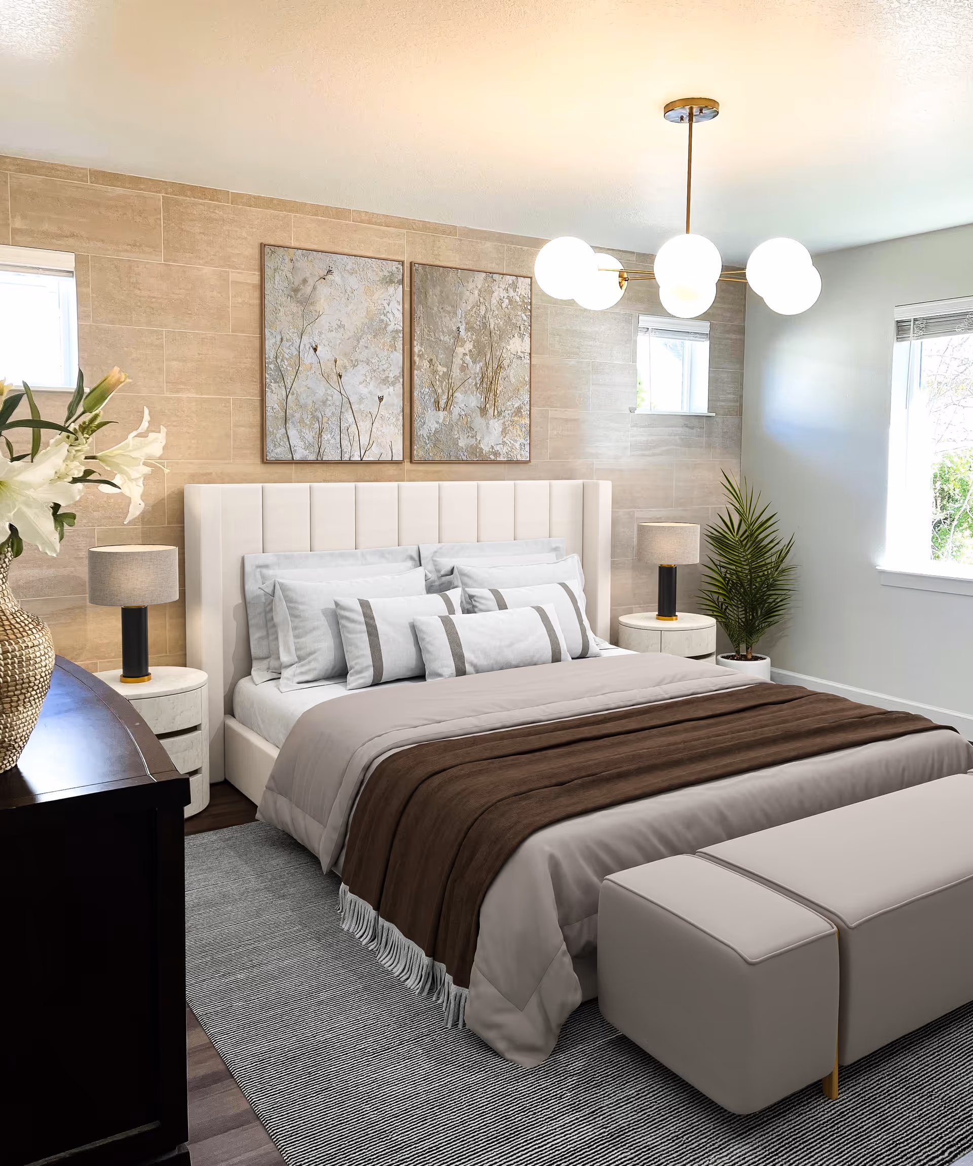 A modern bedroom with a large upholstered bed featuring multiple pillows and a brown throw blanket. The room has two round bedside tables with matching lamps, a dark wooden dresser with a vase of white flowers, two abstract paintings on a beige tiled accent wall, a gray area rug, a beige bench at the foot of the bed, and two windows letting in natural light. A green potted plant is placed near one window.
