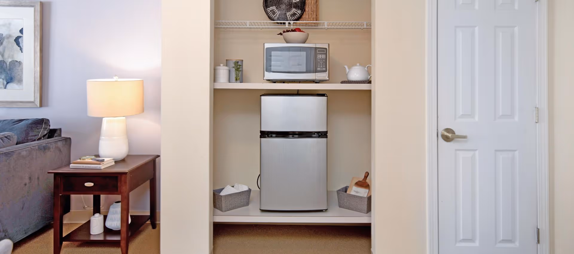 A small kitchenette area with a mini refrigerator and a microwave on a shelf above it. There are two gray baskets on either side of the refrigerator, one containing a cutting board. To the left is a wooden side table with a white lamp and some books, next to a gray couch. A white door is visible on the right side of the image.