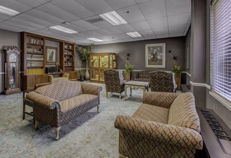 A cozy living room area in a senior living facility featuring two patterned sofas, two floral armchairs, a wooden coffee table, a tall grandfather clock, a wooden bookshelf filled with books, decorative plants, and framed artwork on the walls. The room has a carpeted floor and large windows with blinds allowing natural light to enter.