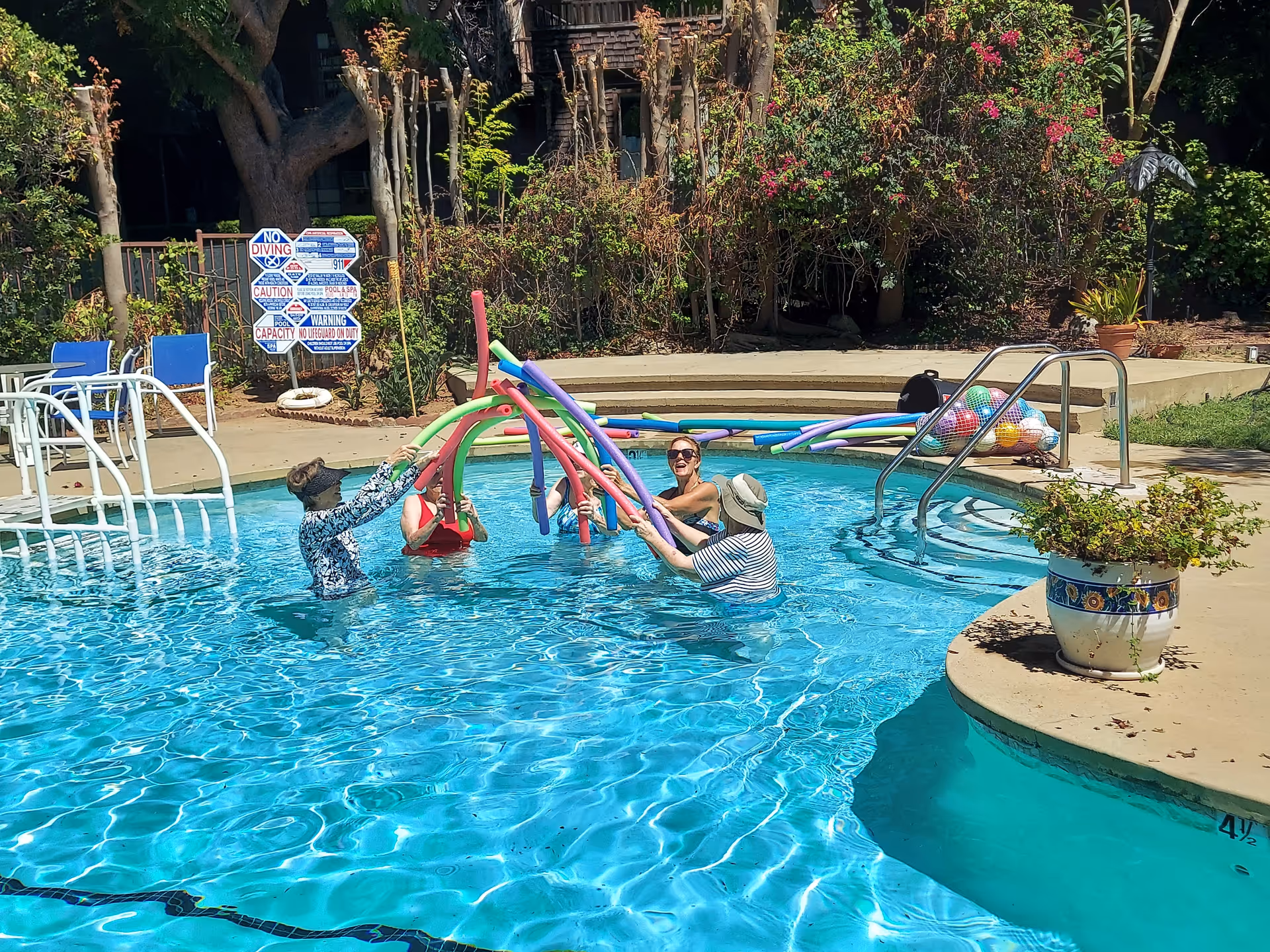 Four people in a swimming pool holding colorful pool noodles above the water, surrounded by greenery and poolside chairs. There is a sign with pool rules in the background and a potted plant on the pool deck.