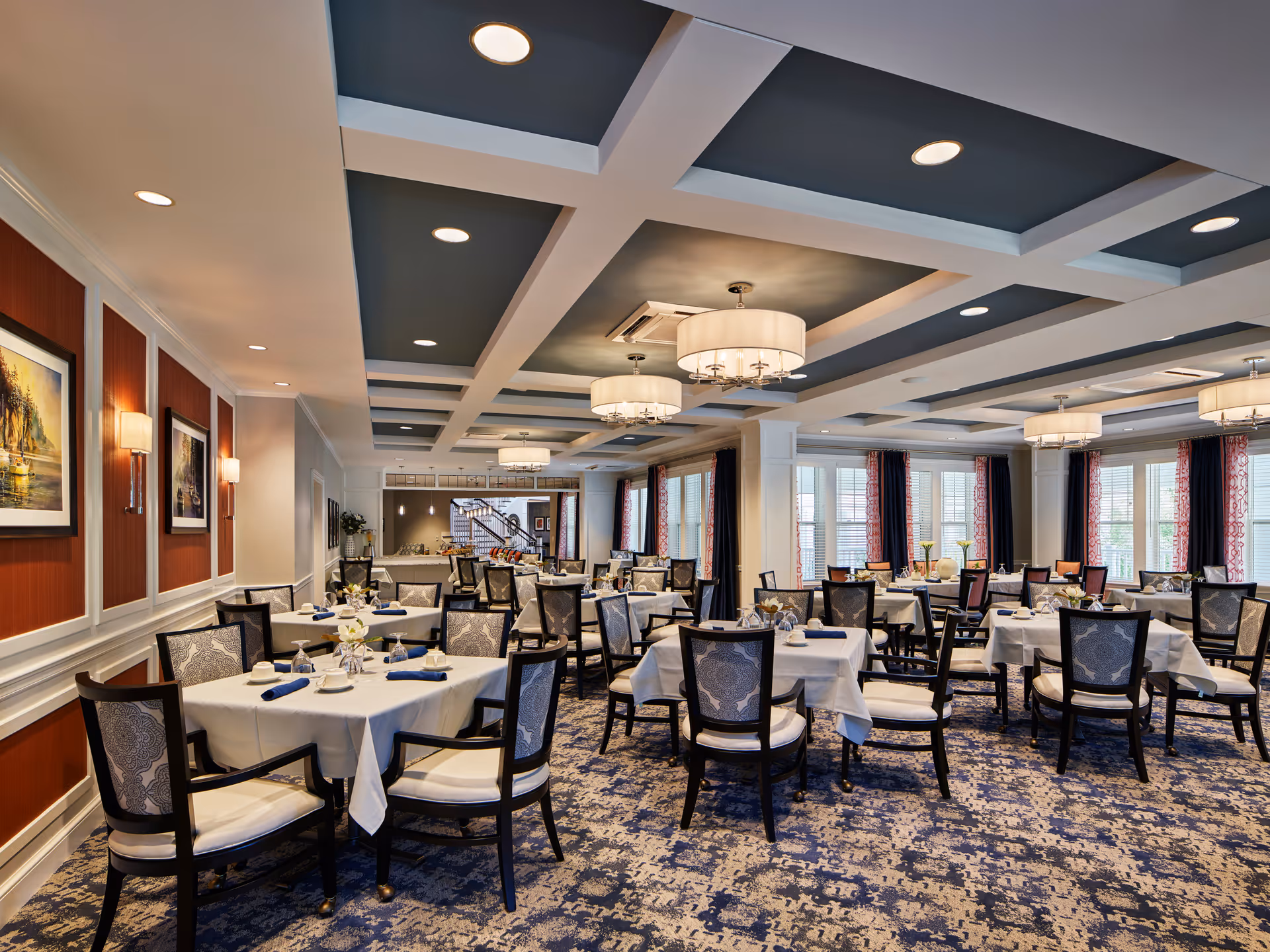 A spacious and elegant dining room with multiple tables covered in white tablecloths, each set with napkins, glasses, and small floral centerpieces. The room features patterned carpet, dark wooden chairs with upholstered backs and seats, large windows with curtains, and a coffered ceiling with modern light fixtures.