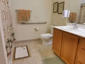 A clean and simple bathroom with a toilet, a sink with wooden cabinets, a mirror, a towel rack with two towels, a shower curtain with a palm tree design, and two bath mats on the tiled floor.