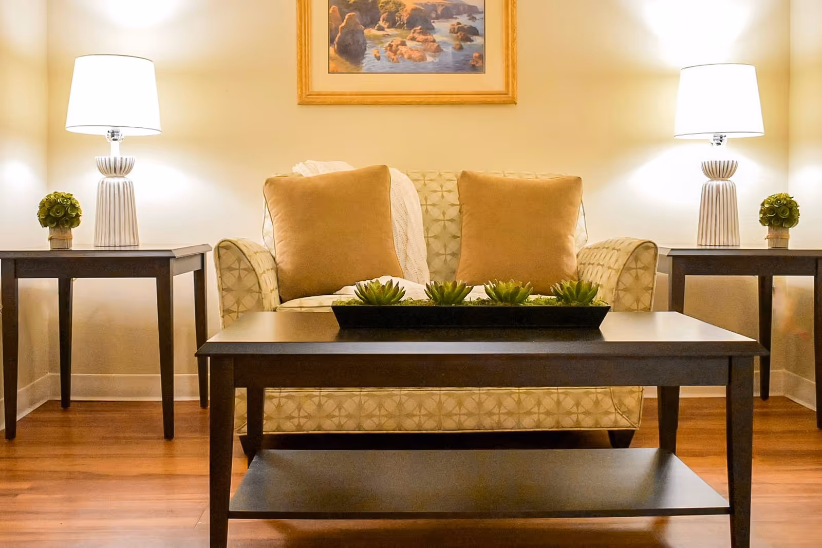 A cozy living room area with a patterned loveseat featuring two brown cushions and a white throw blanket. In front of the loveseat is a dark wooden coffee table with a rectangular planter holding small green succulents. On either side of the loveseat are matching dark wooden side tables, each with a white table lamp and a small green plant. A framed painting of a rocky coastal scene hangs on the wall above the loveseat. The room has warm lighting and wooden flooring.