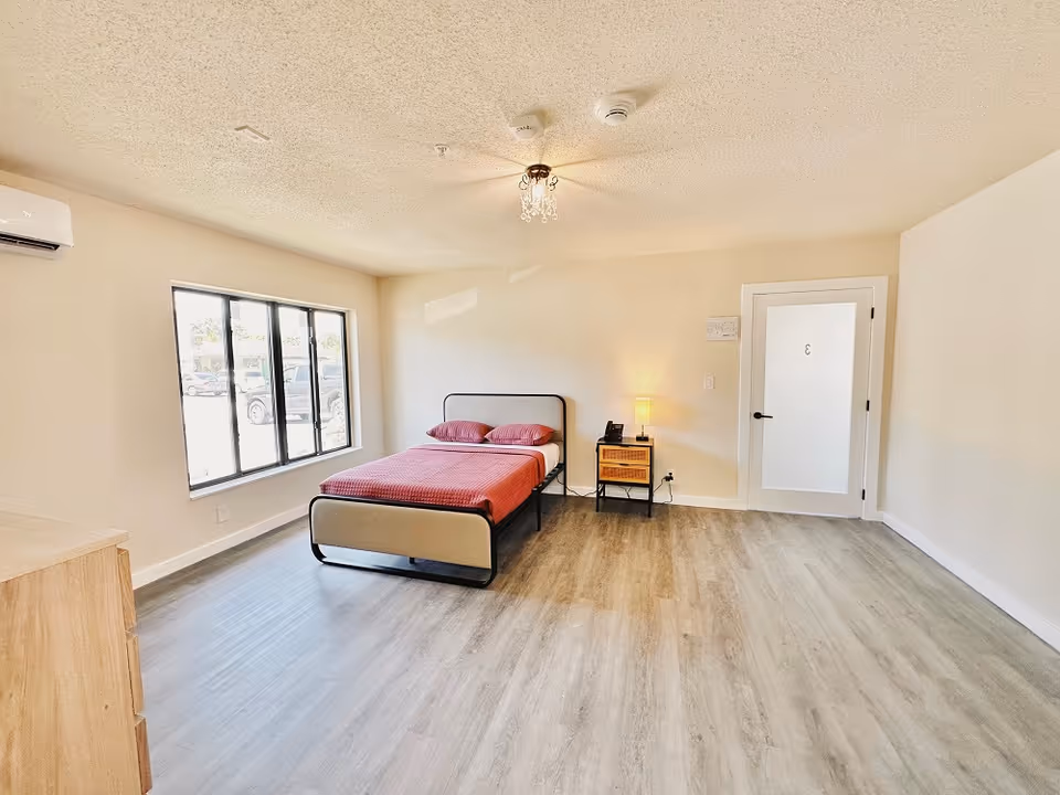A bright, spacious bedroom with light wood flooring, a large window with black frames, a bed with a red bedspread and two pillows, a small nightstand with a lamp and a phone, a wooden dresser, and a door with the number 3 on it.