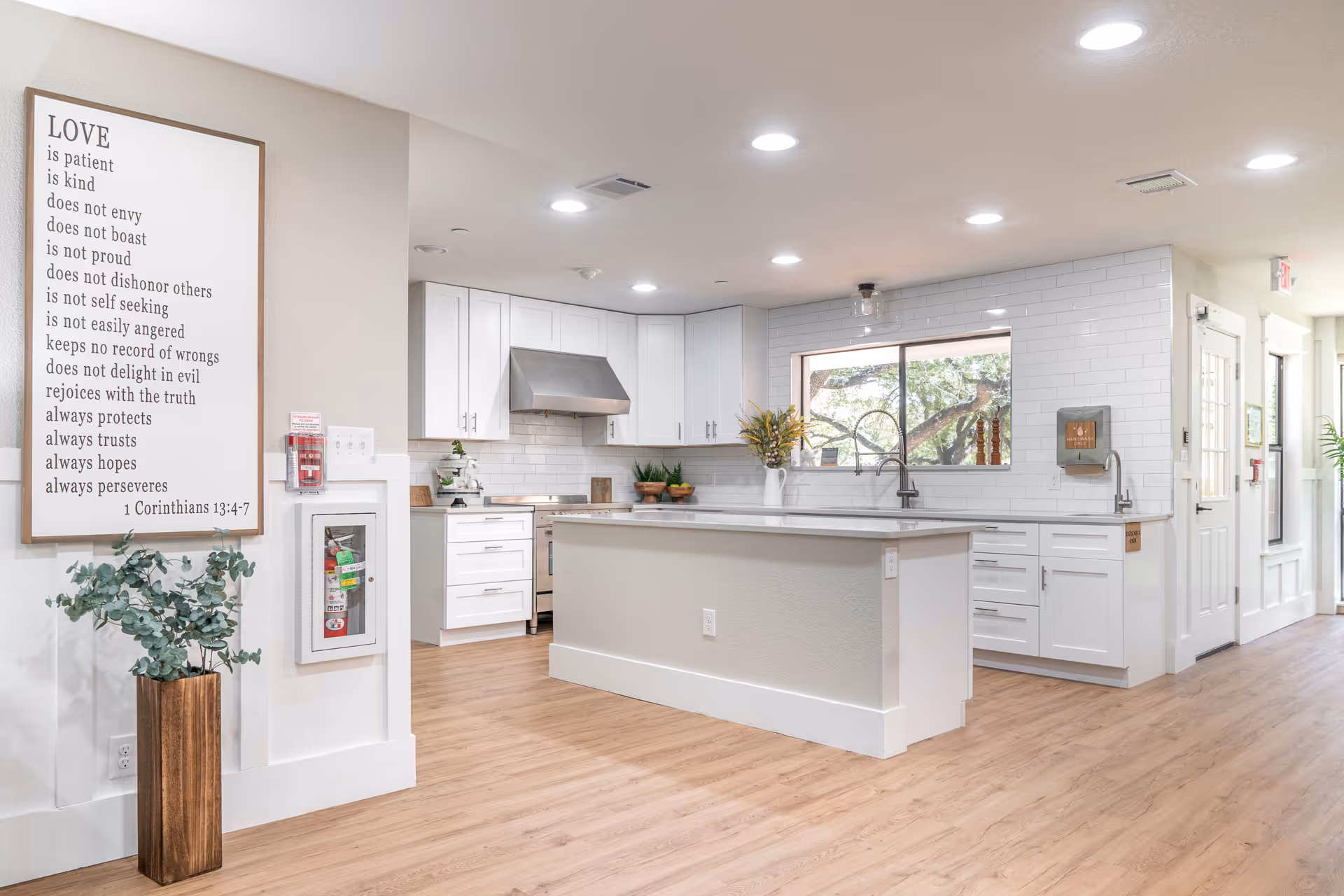 Bright and modern kitchen area with white cabinets, a large island with a light-colored countertop, stainless steel stove and hood, a window above the sink showing trees outside, and wooden flooring. On the left wall, there is a framed sign with a quote from 1 Corinthians 13:4-7 about love, and a tall wooden vase with green foliage. The space is well-lit with recessed ceiling lights.