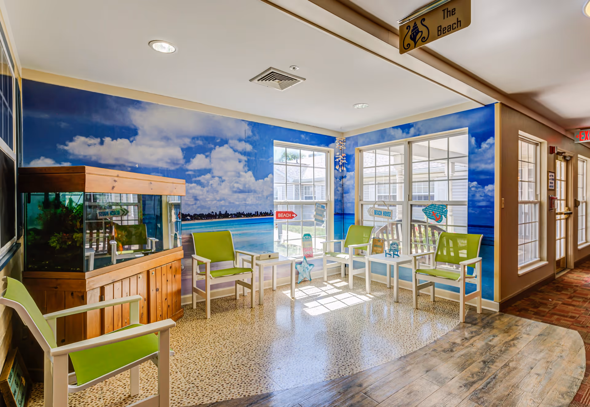 A bright and cheerful seating area with green chairs and small white tables arranged near large windows. The walls are decorated with a beach-themed mural showing a blue sky with clouds and ocean waves. There is a large fish tank with a wooden frame on the left side. A small sign hanging from the ceiling reads 'The Beach'.