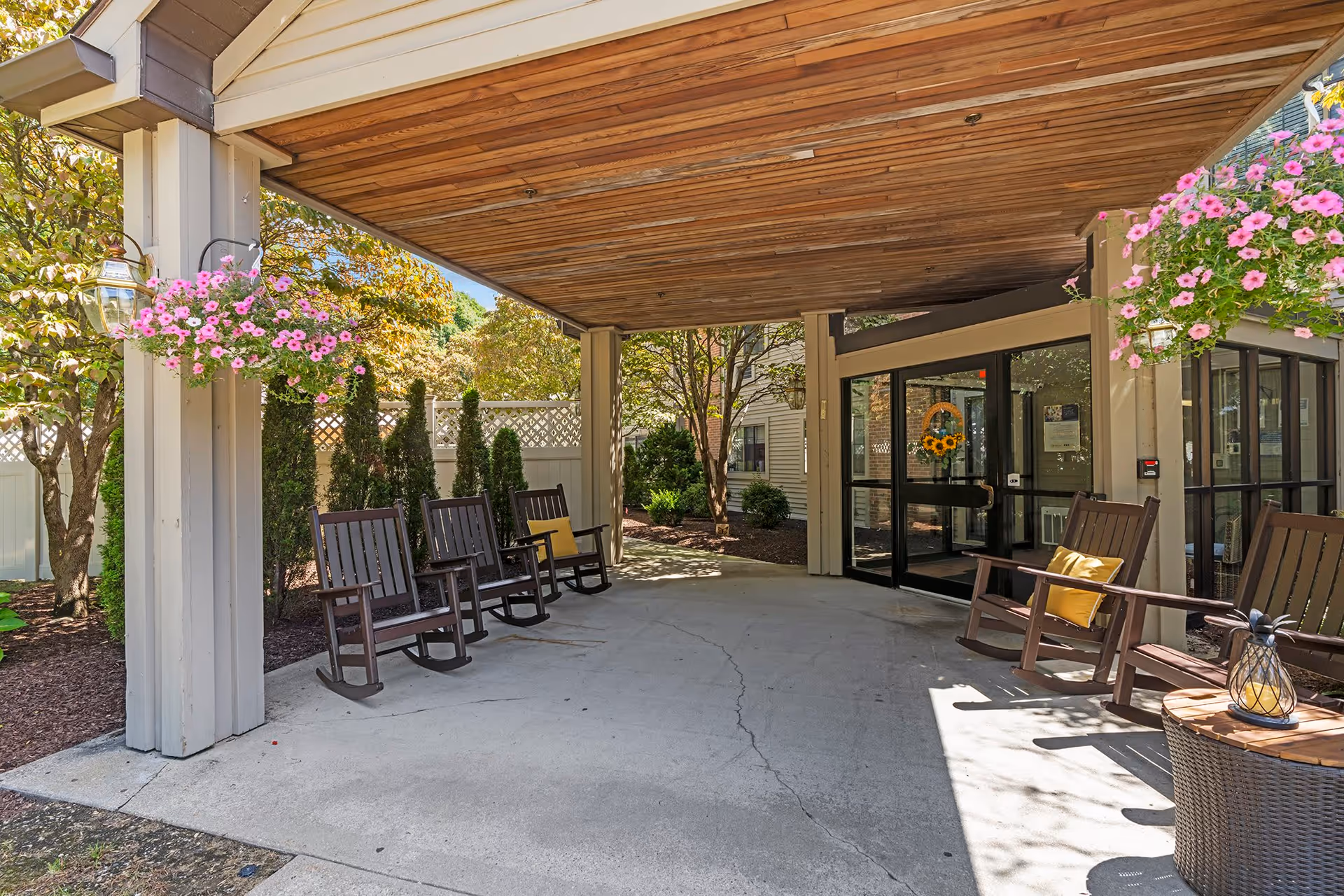 Covered outdoor seating area with wooden rocking chairs and hanging pink flower baskets at the entrance of a building. The area is shaded by a wooden ceiling and surrounded by trees and shrubs.