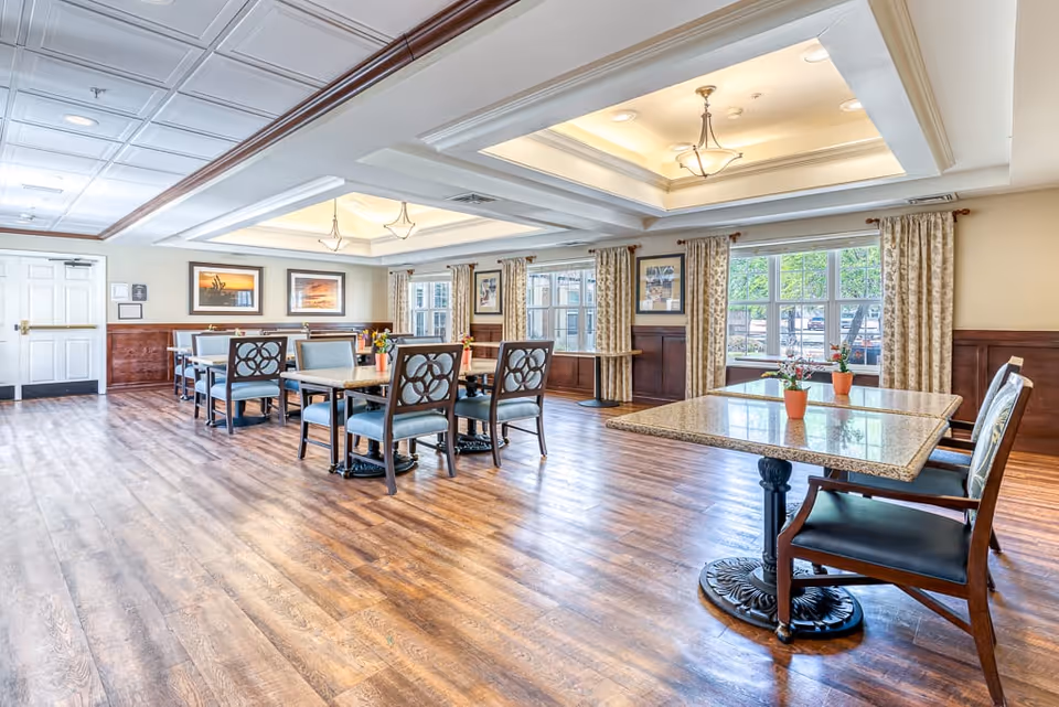 Bright memory care dining room with multiple tables and chairs, hardwood floors, coffered ceilings, and large windows.