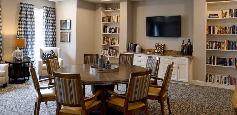 A cozy common area with a round wooden table surrounded by eight striped cushioned chairs. The room features built-in white bookshelves filled with books and decorative items, a flat-screen TV mounted on the wall above a white cabinet, patterned carpet, and a window with blue and white patterned curtains. There are also two armchairs and a side table with a lamp near the window.