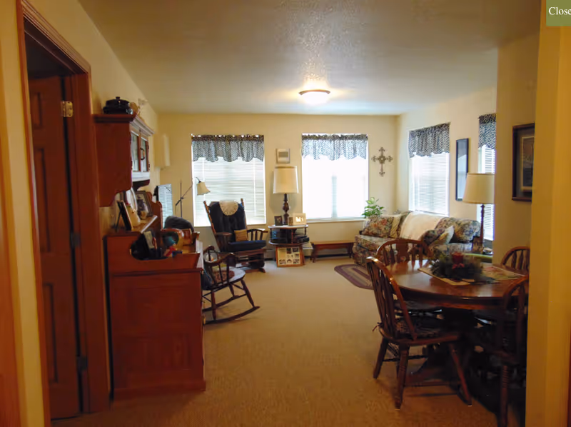 A cozy living room and dining area with a sofa, rocking chairs, a wooden dining table, and three front windows with valances.