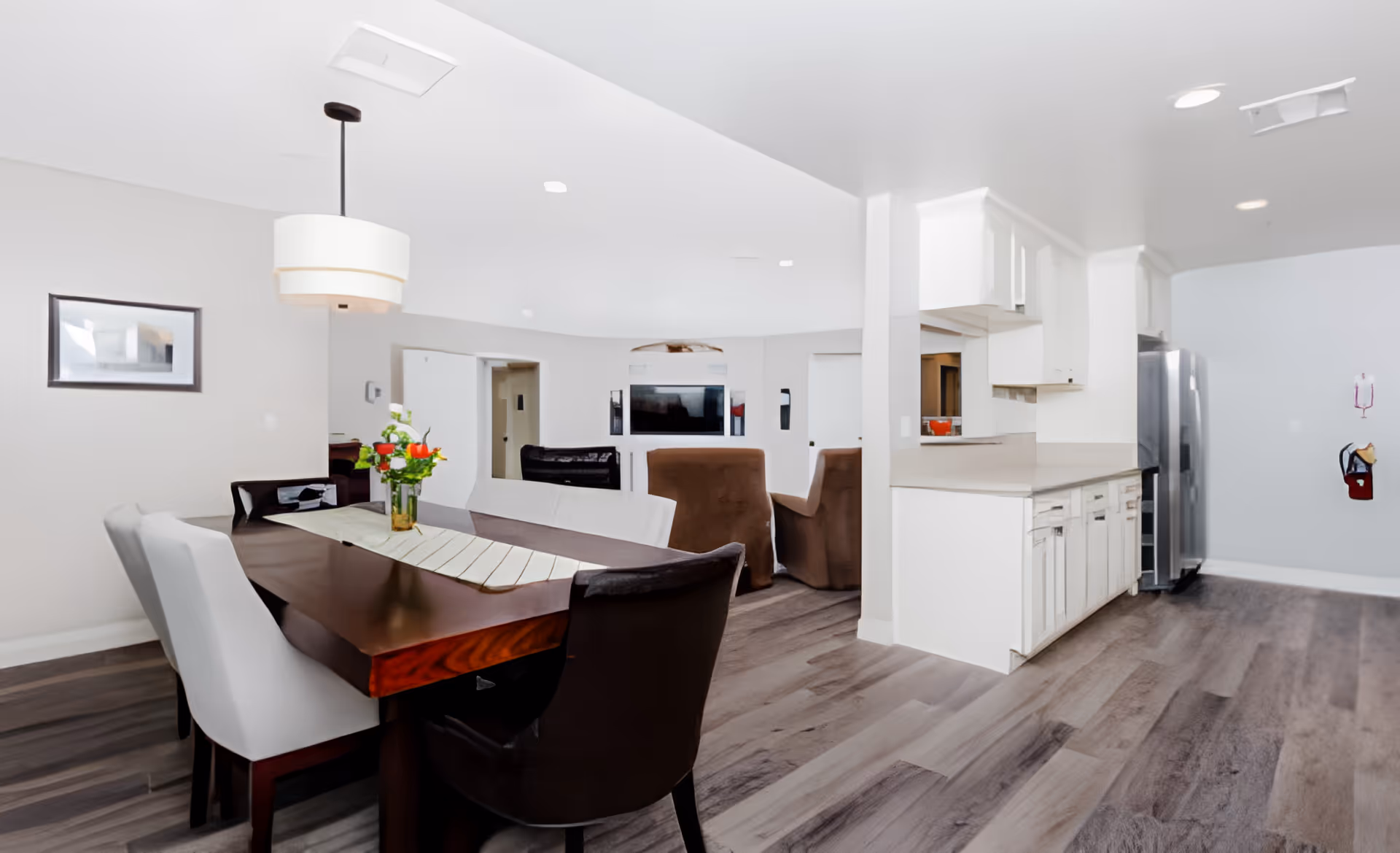 Open-plan dining area with a wooden table and upholstered chairs adjacent to a white kitchen and living area.