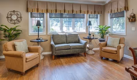 A cozy living room area with two tan armchairs and a green loveseat arranged around two round side tables with lamps. The room has large windows with beige valances, wooden flooring, and decorative plants and wall clock.