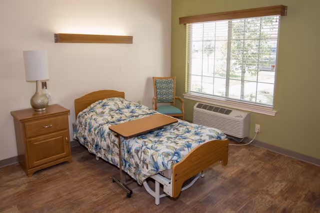 A furnished senior bedroom with an adjustable bed, overbed table, nightstand with lamp, chair, and a window with an air conditioner.