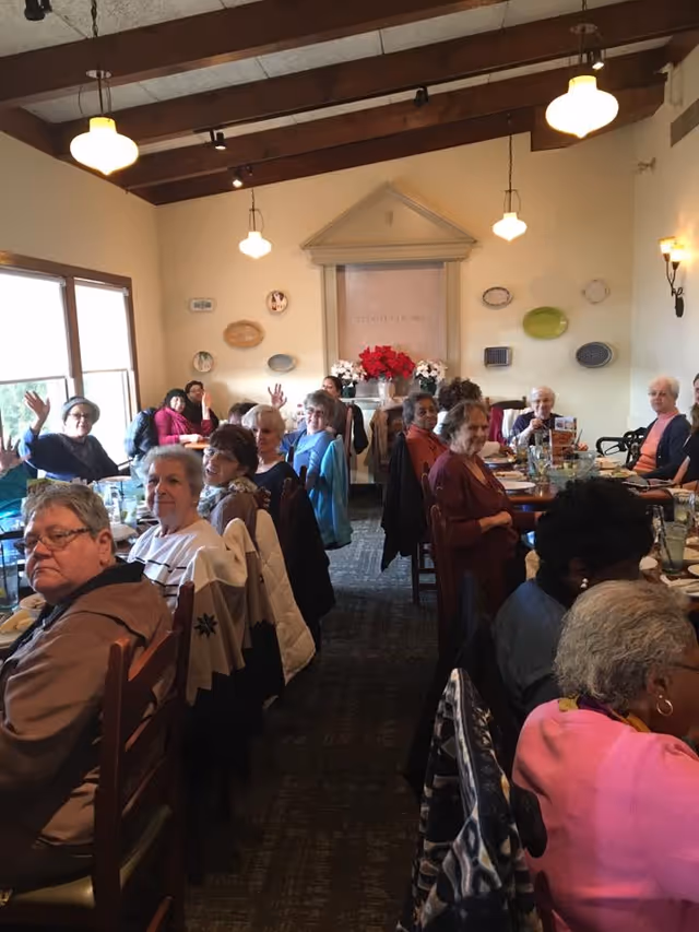 A group of senior residents seated at long tables in a dining room, many looking toward the camera.