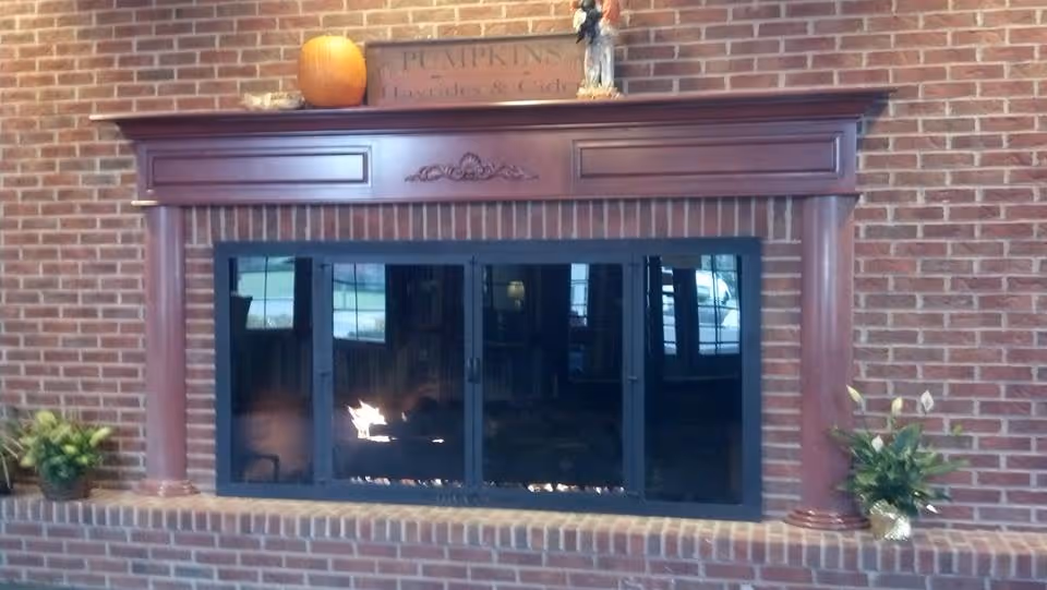 A brick fireplace with a wooden mantel decorated with a pumpkin, a small statue, and a sign that reads 'Pumpkins, Hocuspocus & Cider'. There are two potted plants on either side of the fireplace.
