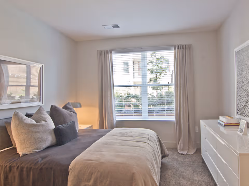 A neatly made bed with multiple pillows in a bedroom featuring a large window with blinds and curtains. There is a white dresser with books and a framed photo on top, and a bedside table with a lamp. The room has light-colored walls and carpeted flooring.