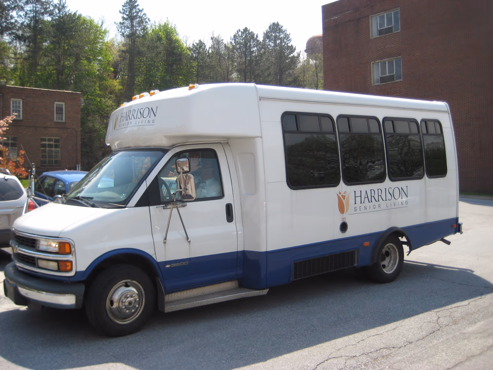A white Harrison Senior Living shuttle bus parked outside near brick buildings and trees.