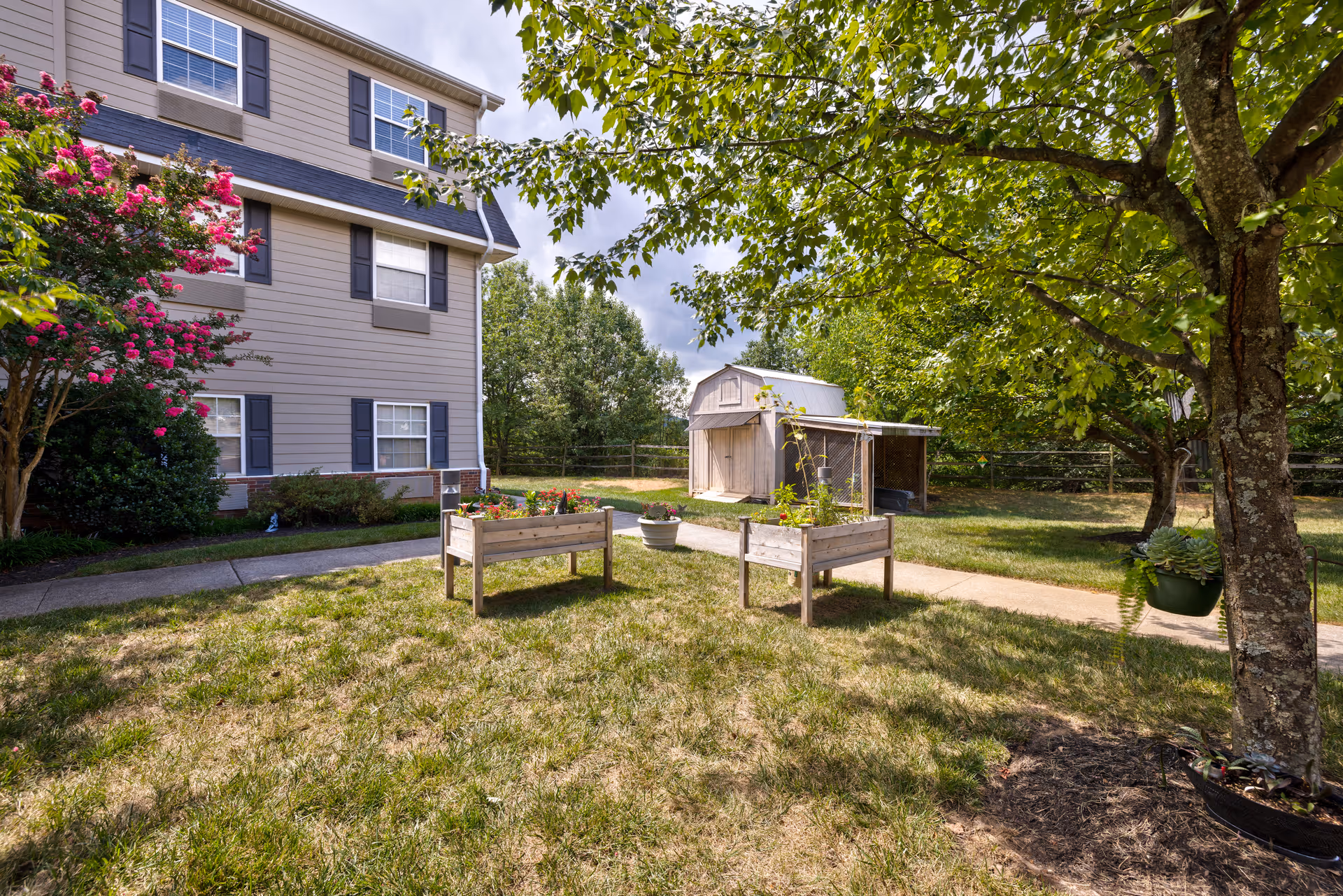 Outdoor garden area at TerraBella Pheasant Ridge featuring a grassy lawn, two raised wooden planter boxes with flowers, a small shed in the background, a tree with hanging potted plants, and part of a multi-story building with windows and black shutters.