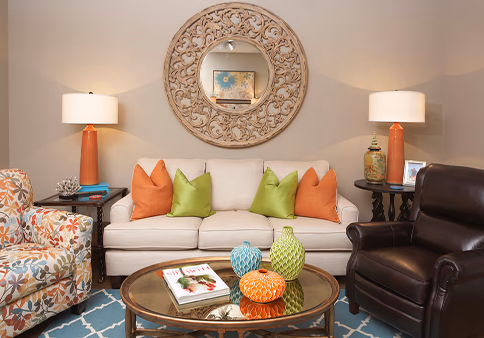 Cozy living room with a beige sofa adorned with orange and green pillows, a patterned armchair and dark leather recliner, a round ornate mirror on the wall, lamps on side tables, and a glass-top coffee table with colorful vases.