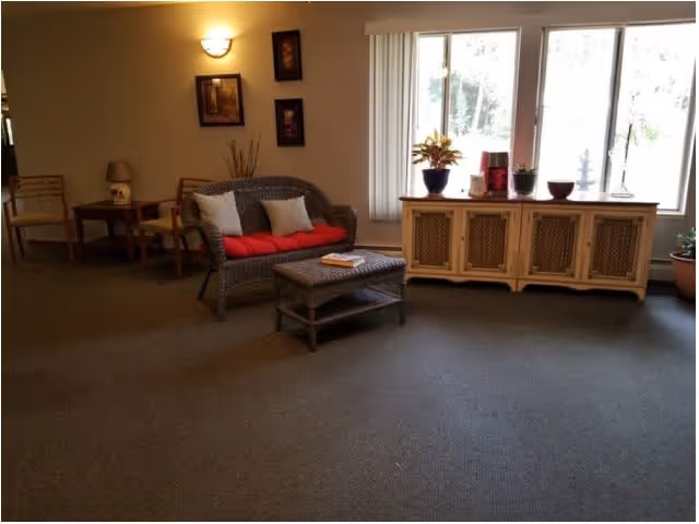 A cozy living room area with a wicker loveseat featuring red and white cushions, a matching wicker coffee table with a book on it, a wooden sideboard with plants and decorative items on top, and a large window with vertical blinds letting in natural light. There are framed pictures on the wall and a small table with chairs in the background.