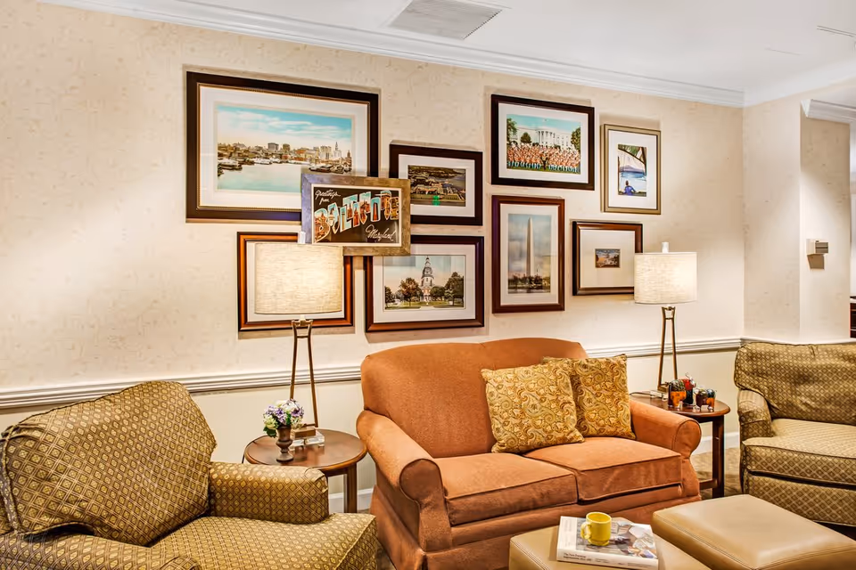 A cozy living room area with a brown sofa adorned with patterned cushions, two patterned armchairs, two side tables each with a lamp, and a wall decorated with framed pictures and artwork.