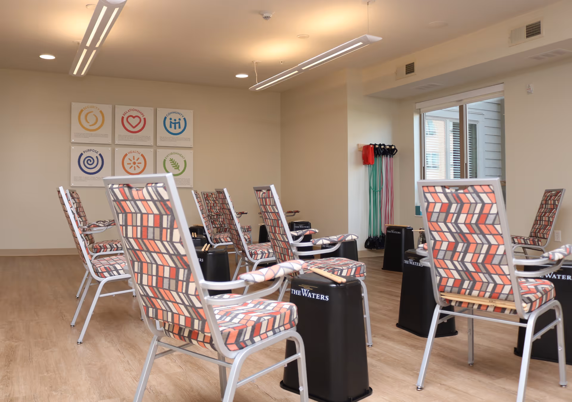 A bright room with several patterned chairs arranged in a circle, each paired with a black drum labeled 'The Waters'. Resistance bands hang on the wall near a window, and colorful motivational posters are displayed on the far wall.