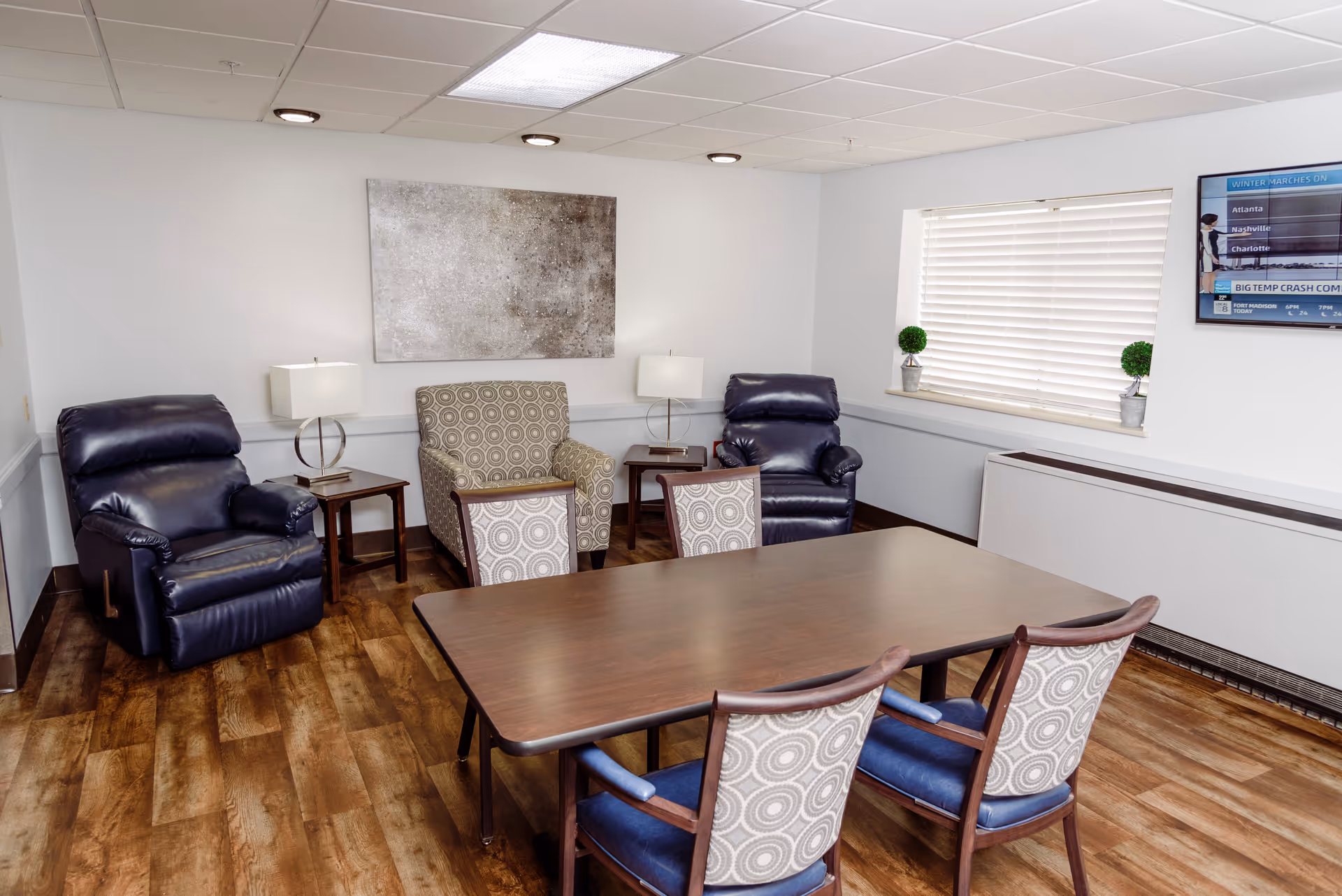 Well-lit common room with a wooden table, upholstered chairs, two leather recliners, side tables and a wall-mounted TV.