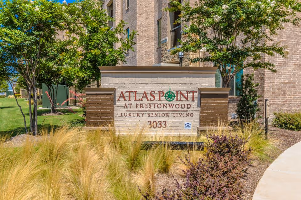 Outdoor view of the entrance sign for Atlas Point at Prestonwood, a luxury senior living facility, surrounded by landscaping with ornamental grasses, shrubs, and trees, with a brick building in the background.
