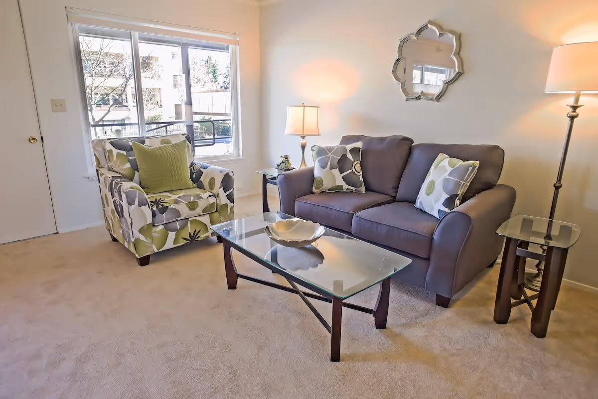 A cozy living room with a gray sofa adorned with floral patterned pillows, a matching armchair with a green cushion, a glass coffee table with a decorative bowl, two side tables each with a lamp, a decorative mirror on the wall, and a large window letting in natural light.