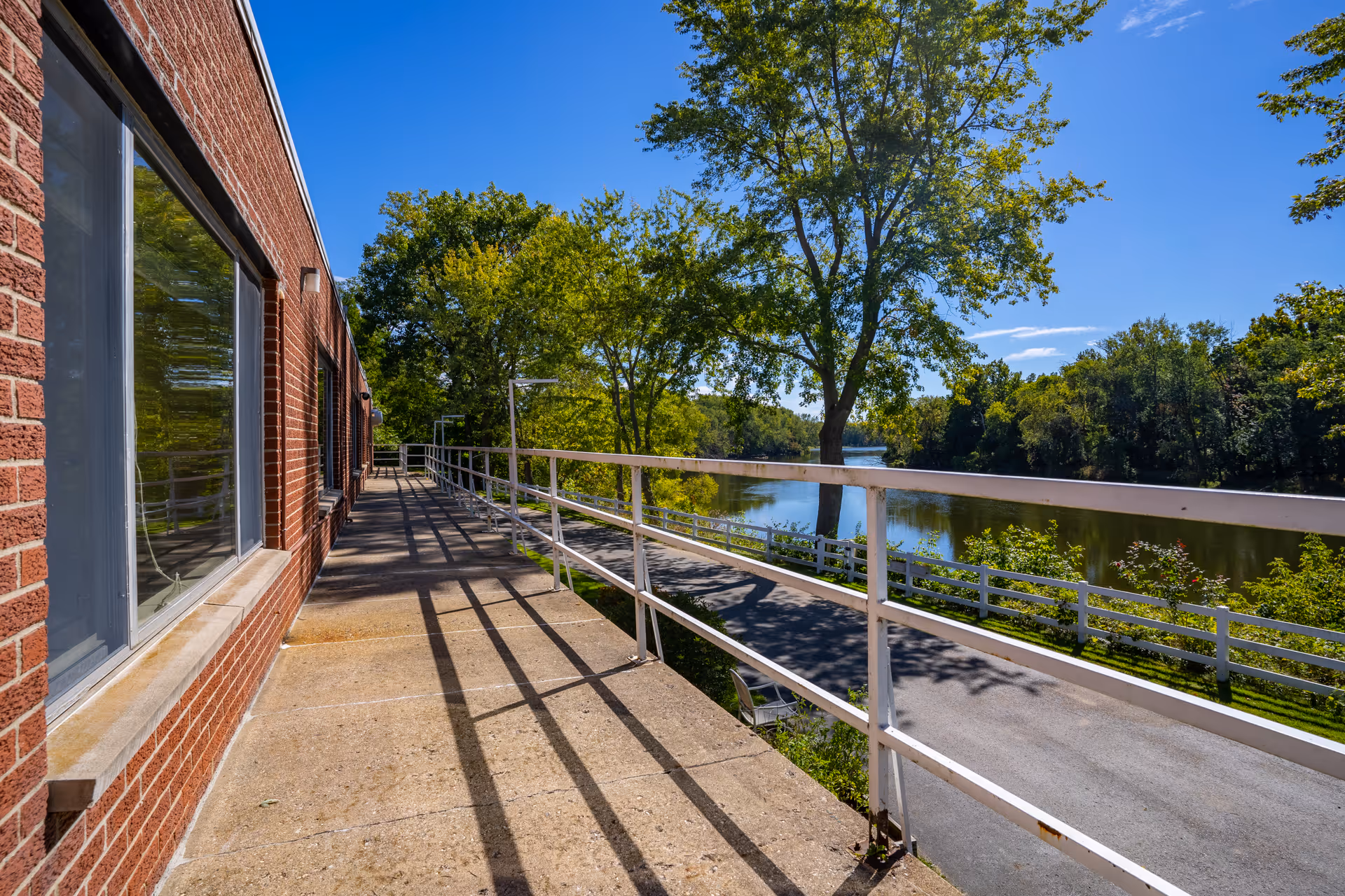 Outdoor walkway alongside a brick building with large windows, overlooking a river with trees and a clear blue sky.