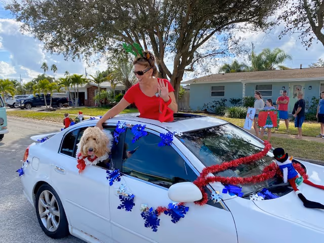 A white car decorated with blue snowflake garlands and red tinsel is driving down a residential street. A woman wearing a red shirt and reindeer antlers is leaning out of the sunroof waving, and a fluffy dog with a red bow is sticking its head out of the rear passenger window. People are standing on the sidewalk in the background, and there are houses and trees along the street.