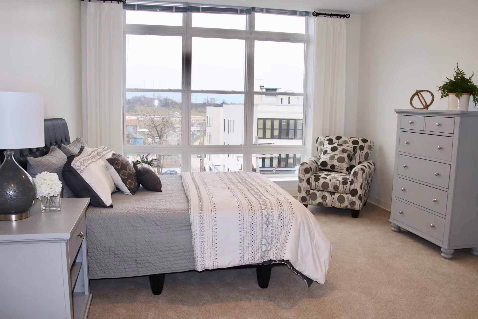 Bright modern bedroom with a large window, a made bed with pillows, a patterned armchair, and a gray dresser.