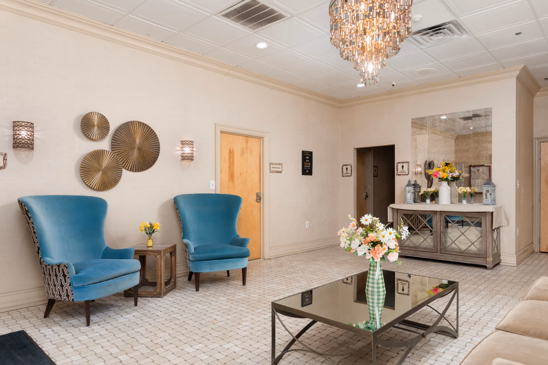 A well-lit interior lounge area with two blue upholstered armchairs, a small wooden side table with a yellow flower vase, a mirrored console table with floral arrangements, and a glass coffee table with a green checkered vase holding a bouquet of flowers. The room features beige walls, tiled flooring, decorative wall sconces, and a chandelier hanging from the ceiling.