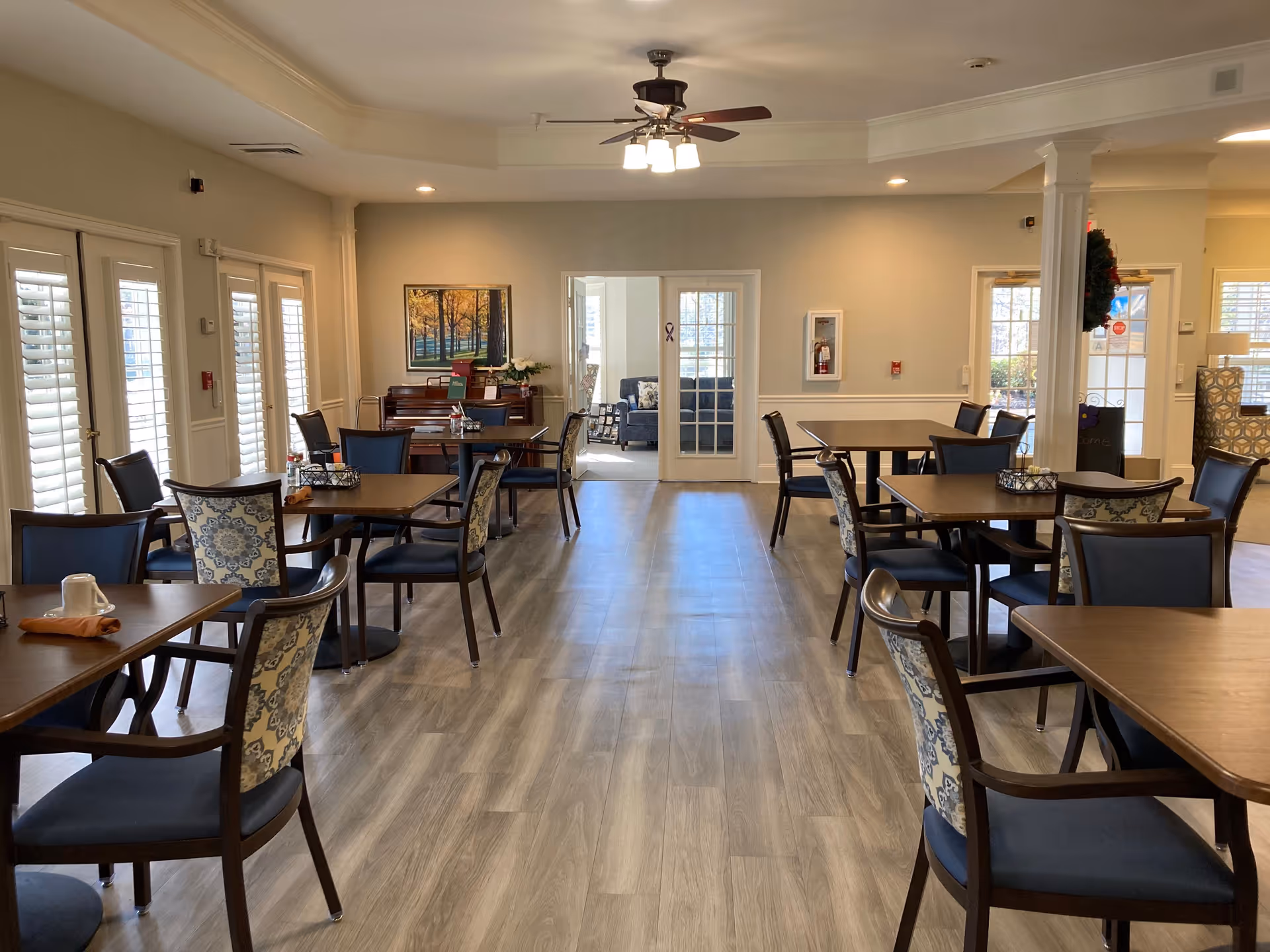 A bright dining room in a senior living facility with several wooden tables and chairs arranged neatly. The chairs have blue and patterned upholstery. The room has light-colored walls, wood flooring, and a ceiling fan with lights. French doors lead to another room with a sofa, and there is a piano against the far wall with a framed picture above it. Large windows with white shutters allow natural light to fill the space.